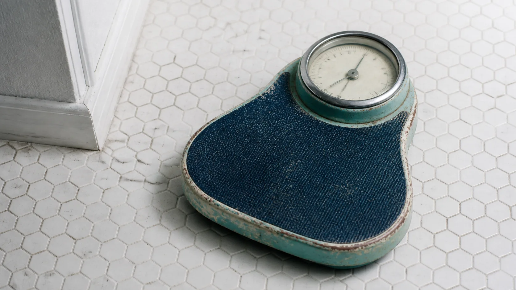 Old mechanical bathroom scale with a round dial standing on white hexagonal floor tiles, worn enamel, soft overhead light