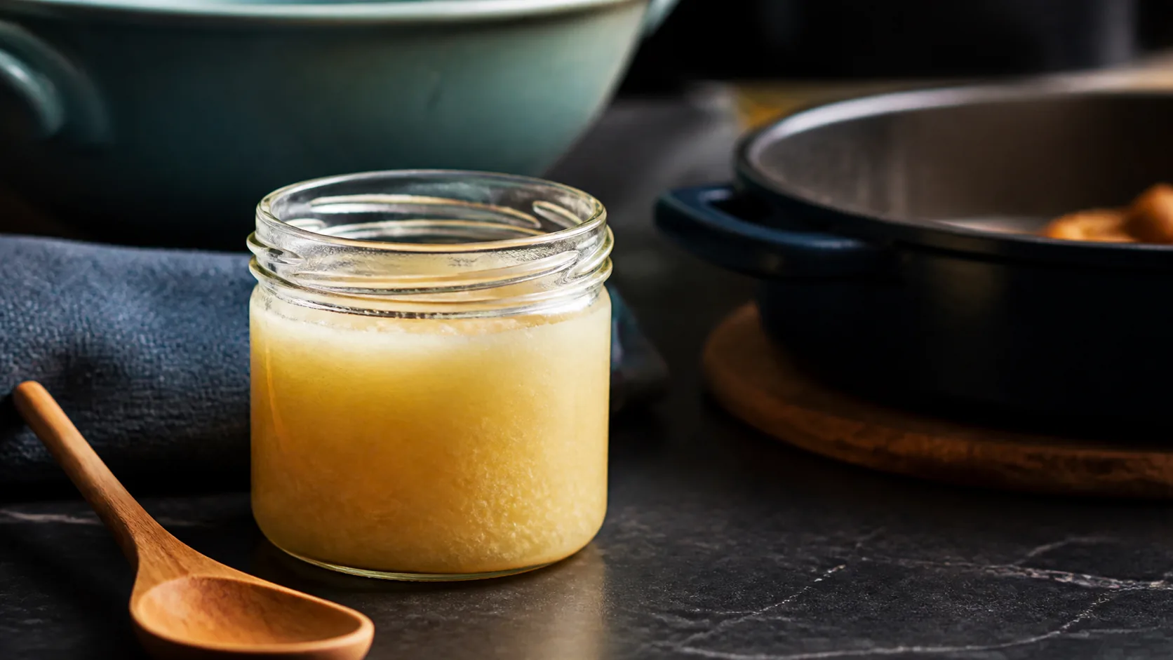 Small glass jar of pale golden rendered fat solidifying on a dark kitchen counter beside a wooden spoon, warm side light