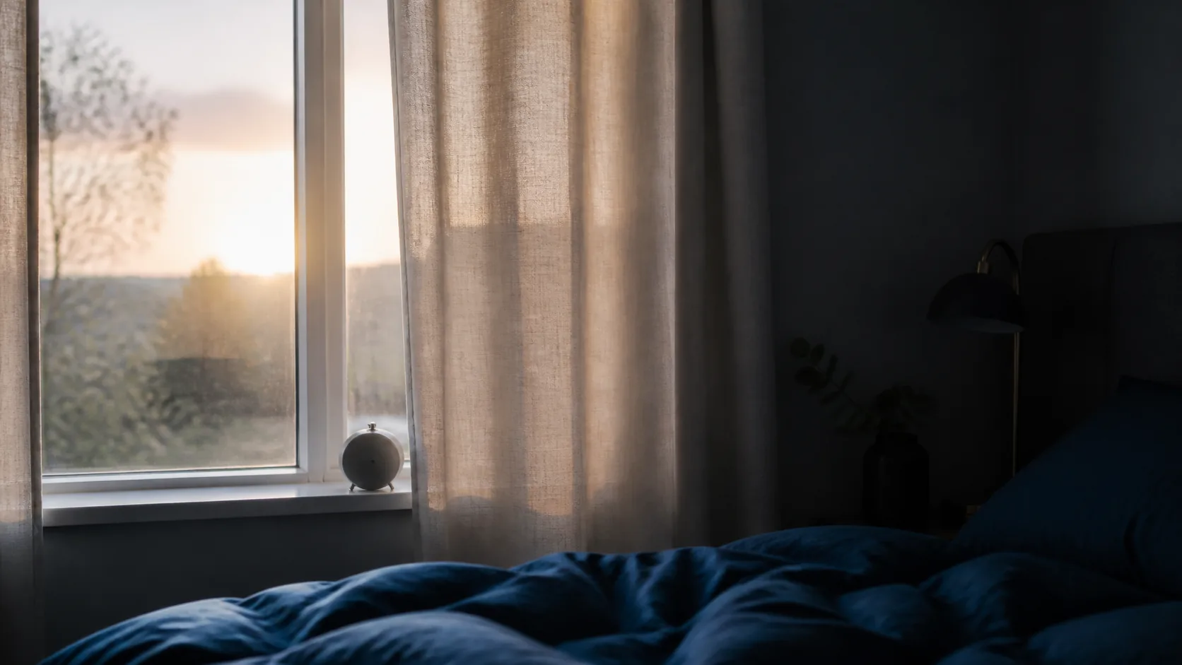 Bedroom window at first light with pale linen curtains half drawn, the room split between night shadow and dawn