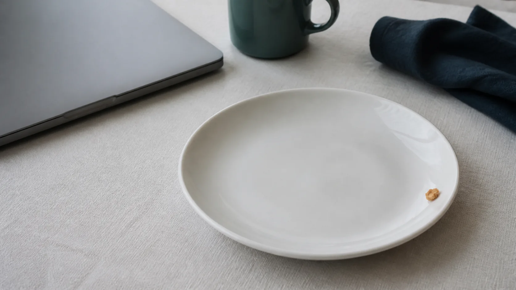 Empty white ceramic plate on a pale linen tablecloth beside a closed laptop, a single crumb of biscuit at the rim