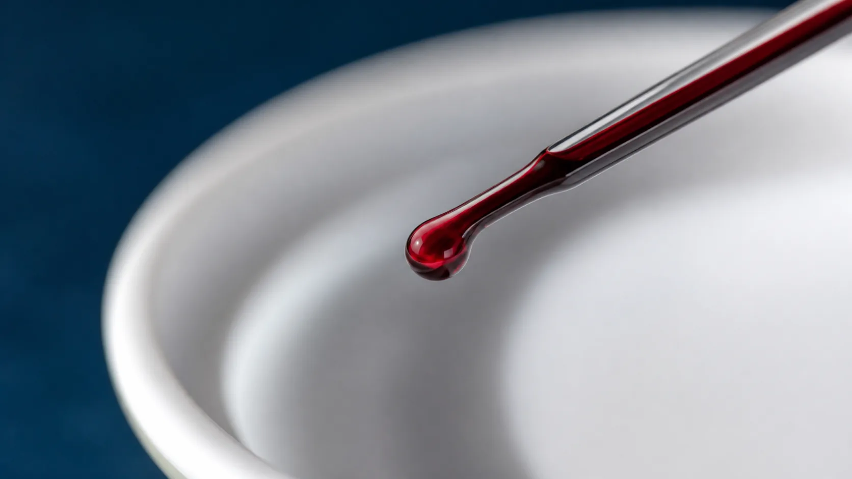 Deep red drop of liquid suspended on the tip of a glass rod above a white dish, extreme macro, hard side light