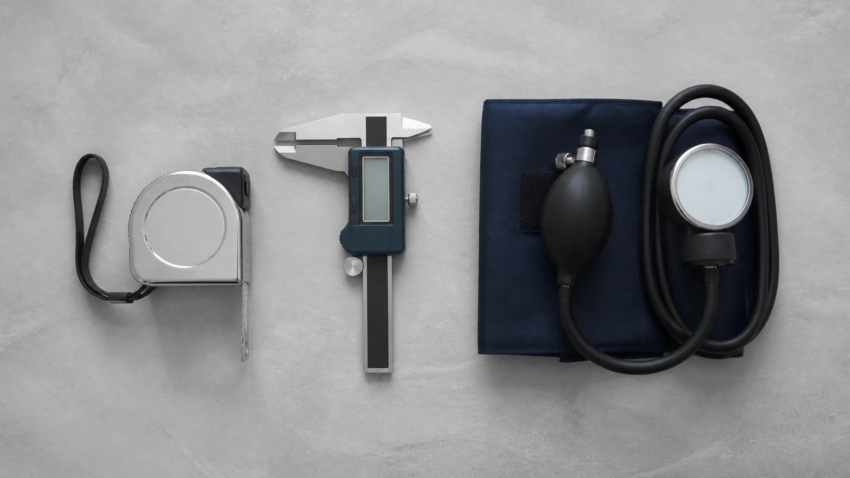 Steel tape measure, a digital caliper and a blood-pressure cuff laid out in a neat row on a pale grey work surface
