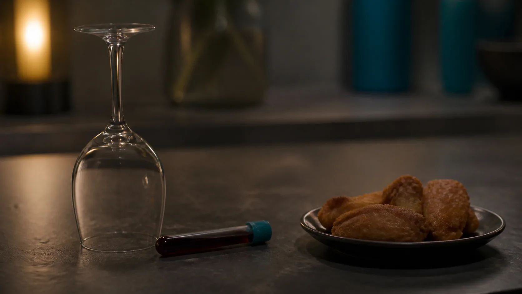 Empty wine glass standing upside down on a dark slate counter beside a capped blood tube and a small plate of fried food g...