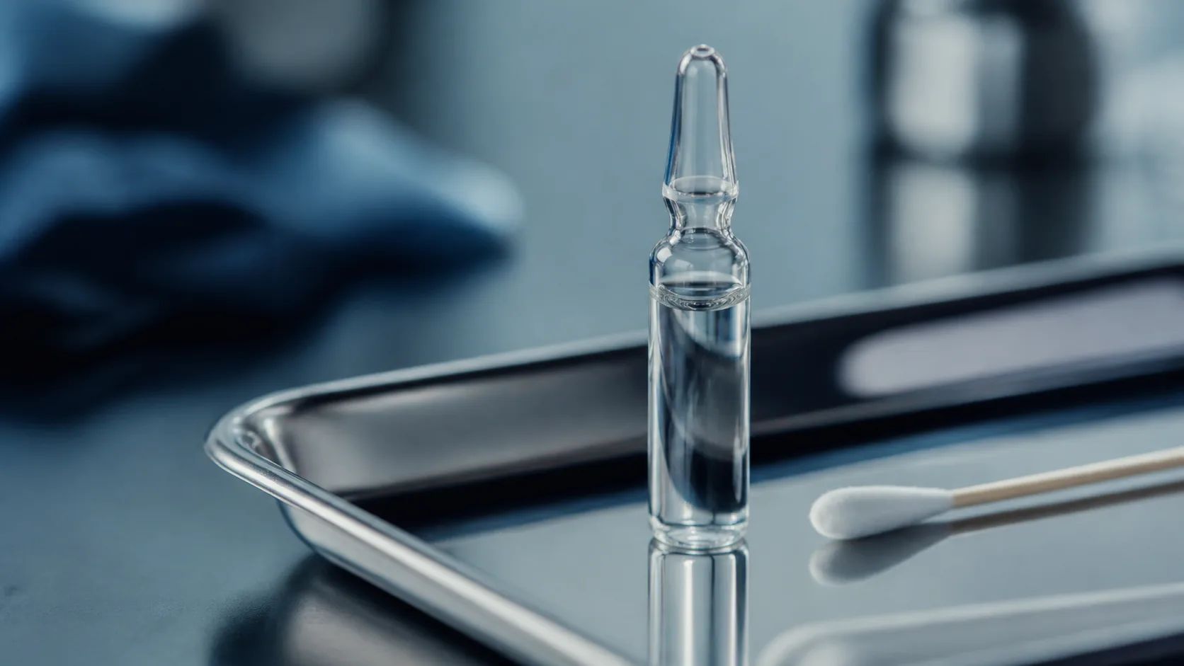 Small clear glass ampoule standing upright on a stainless steel tray beside a cotton swab, cold clinical light