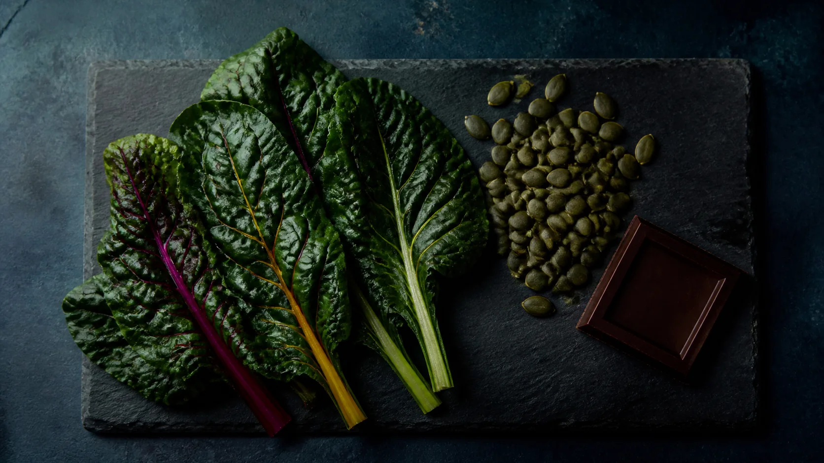 Dark green chard leaves, pumpkin seeds and a square of dark chocolate arranged on a slate board, top-down