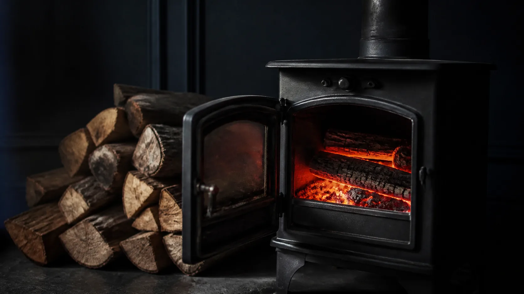 Wood-burning stove with its door open showing hot embers, a stack of split logs beside it, dark room, warm firelight