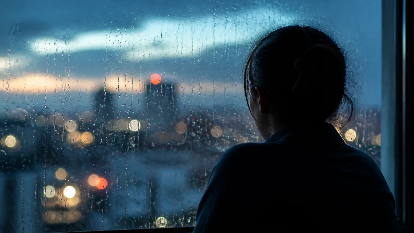 Person's silhouette at a rain-streaked window at dusk, shoulders drawn up, city lights blurred beyond the glass