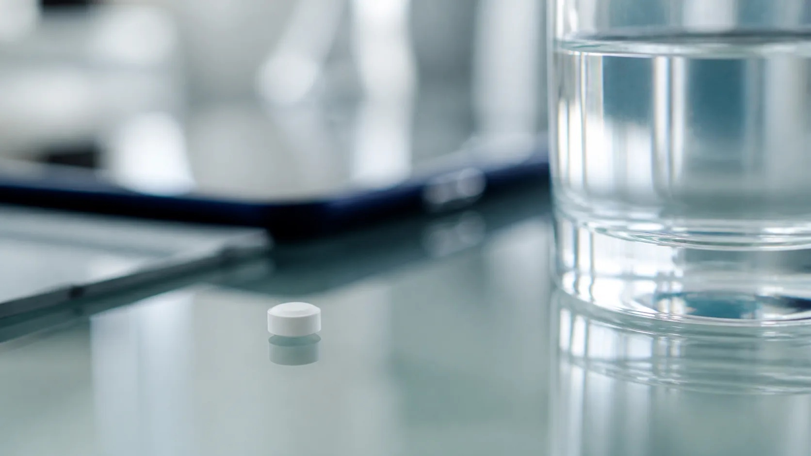 Single small white tablet on a clean glass surface beside a glass of water, extreme macro, cold clinical light
