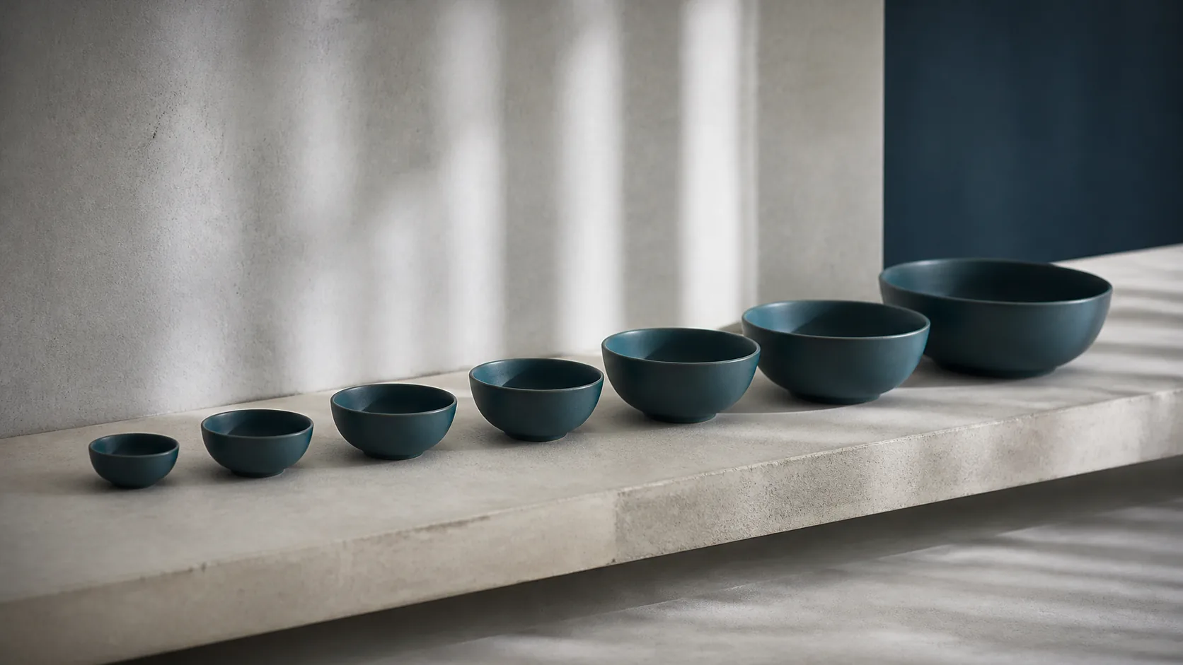 Row of seven identical unlabeled ceramic bowls of graduating size on a long pale concrete shelf