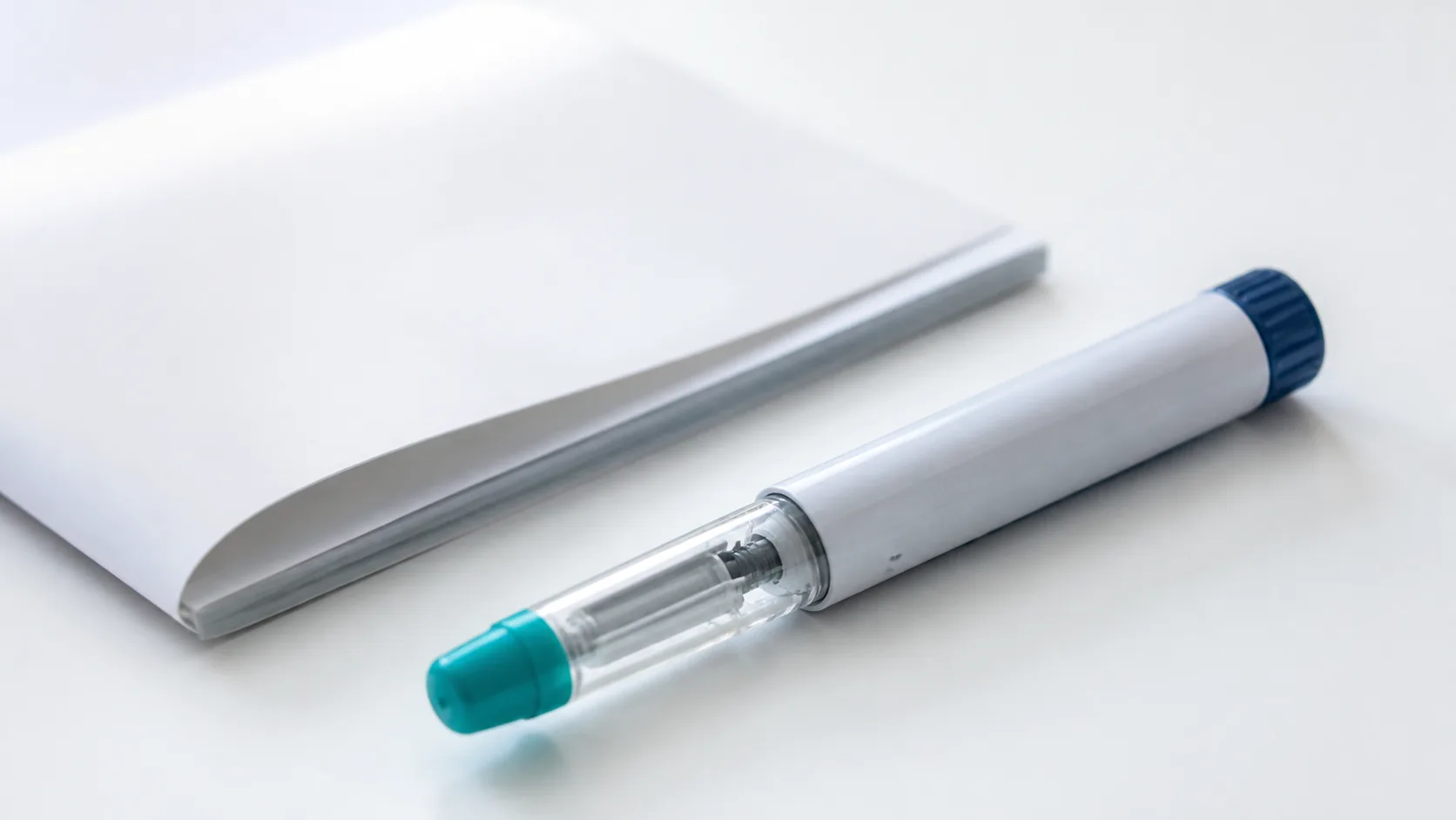 Single unbranded medical pen injector lying on a clean white surface beside a folded prescription pad, cool even light