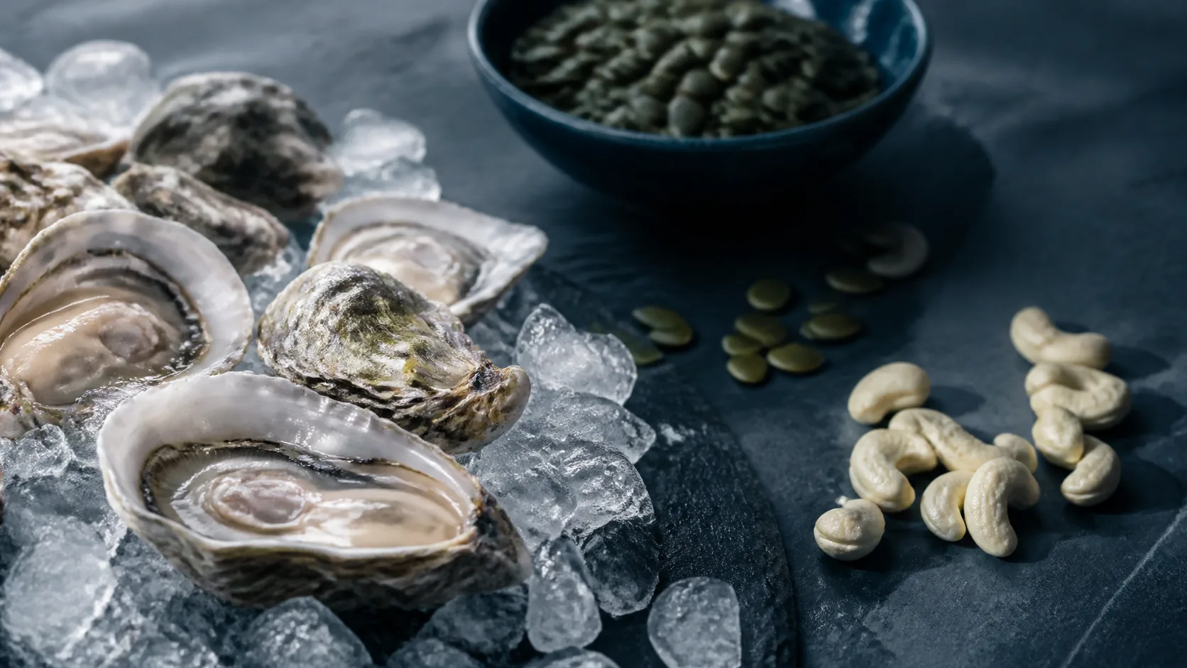 Handful of oysters on crushed ice beside pumpkin seeds and a cashew scatter on dark slate, cold light with a pearl sheen